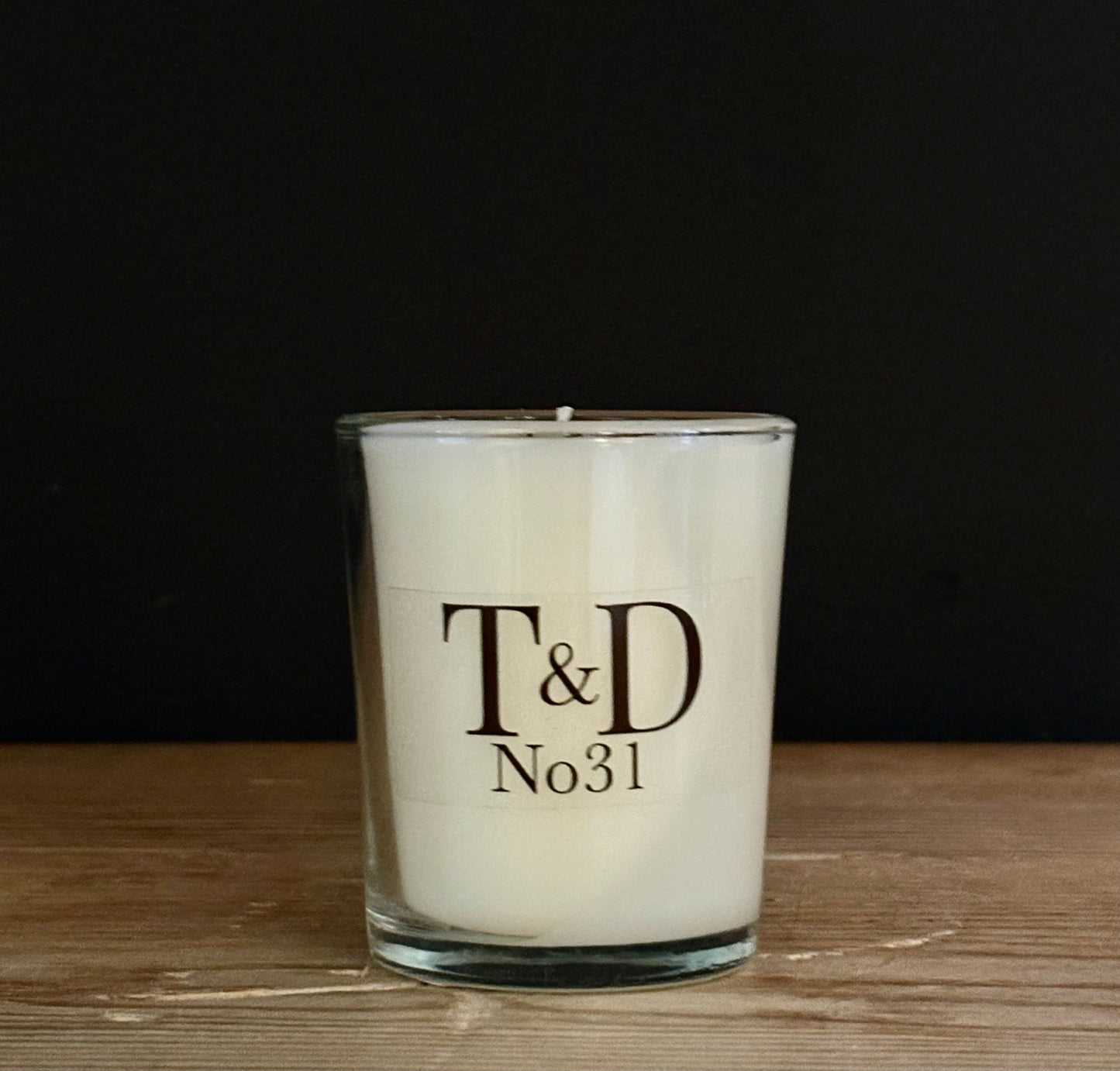 A white scented candle in a clear glass jar with a black printed logo reading 'T&D No31', set on a wooden surface against a dark background.