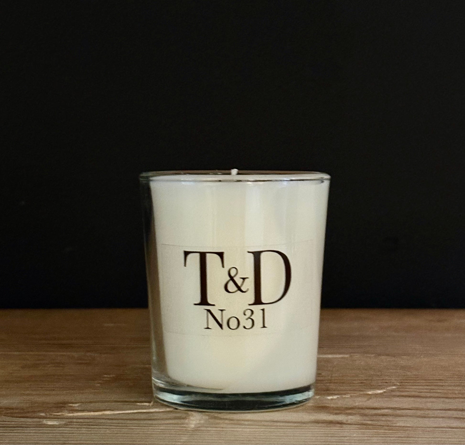 A white scented candle in a clear glass jar with a black printed logo reading 'T&D No31', set on a wooden surface against a dark background.