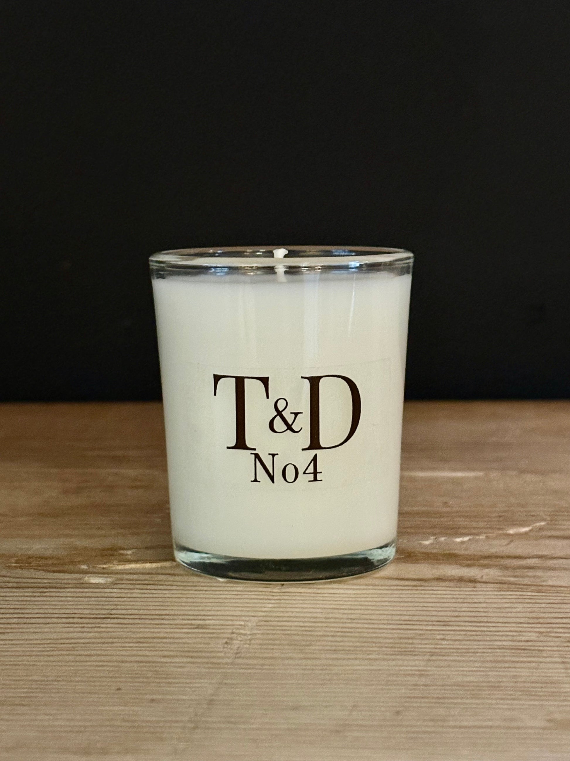 A white soy wax candle in a clear glass jar with a wick, featuring the text 'T&D No4' printed in black on the front, set on a light wooden surface against a dark background.