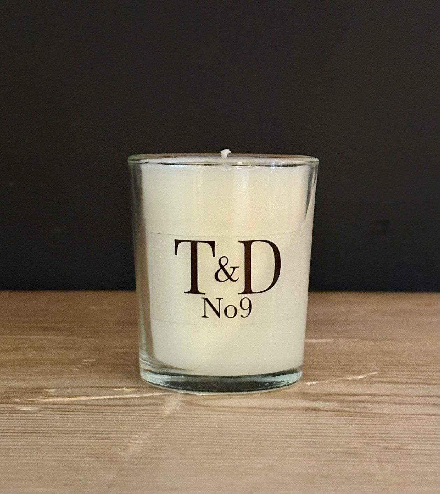 A white wax candle in a clear glass jar with the text 'T&D No9' printed on the front, set on a light wood surface against a dark background.