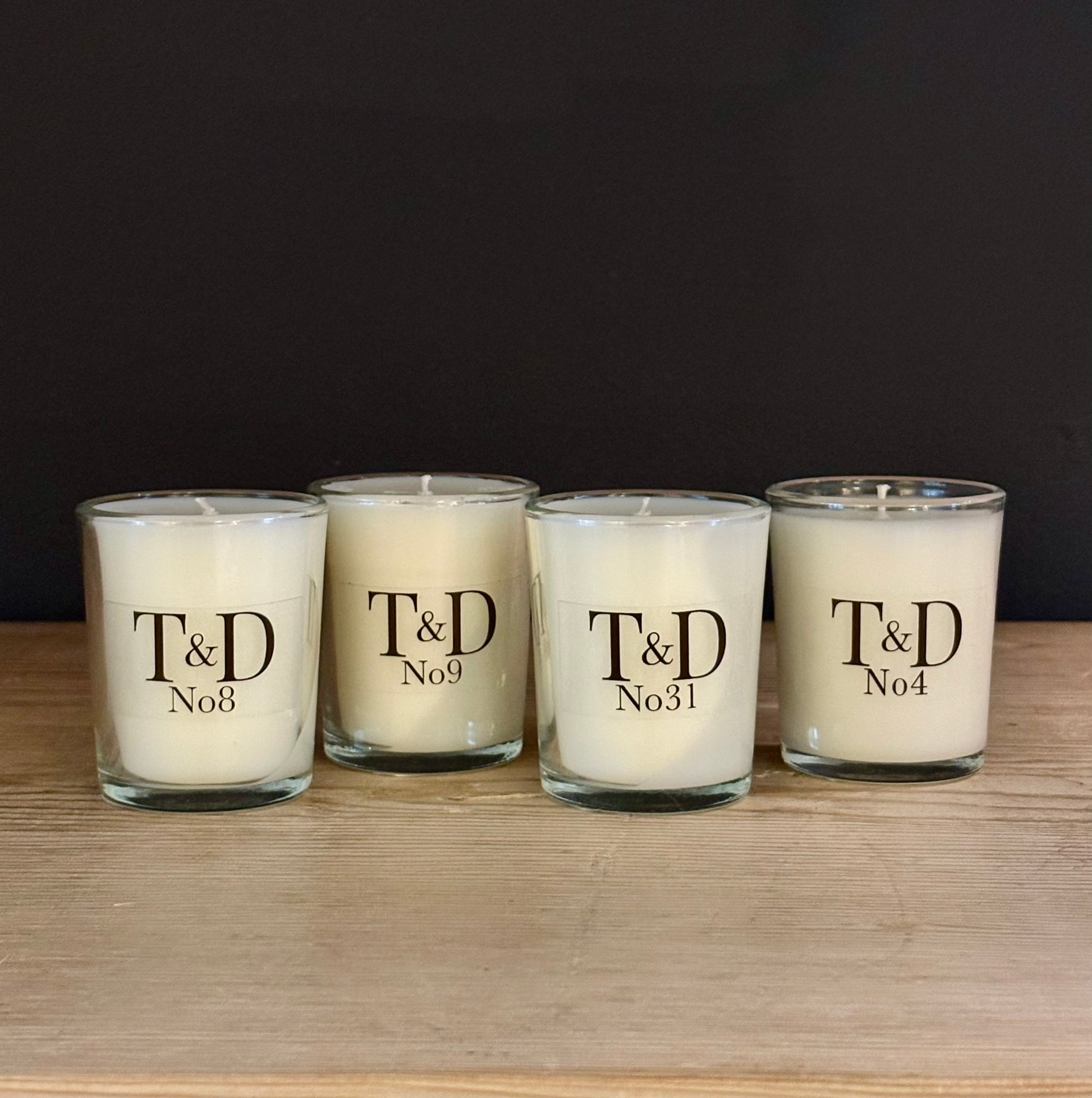 Four white scented candles in clear glass jars with black printed text reading 'T&D No4', 'T&D No8', 'T&D No9', and 'T&D No31', arranged on a light wood surface against a dark background.