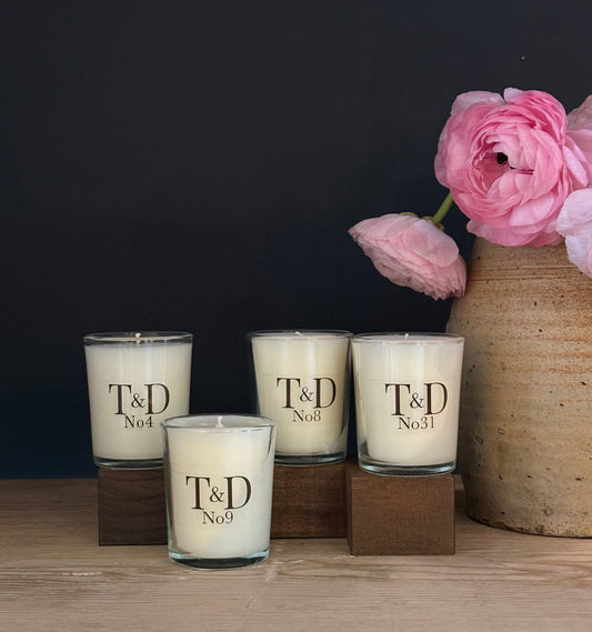 Four T&D scented candles in clear glass holders with dark wood bases, arranged on a light wood surface against a dark navy background, with pink flowers in a ceramic vase to the right.