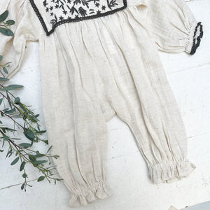 off white linen baby jumpsuit with black embroidery on chest and black trim on end of sleeves