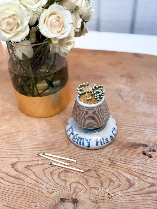 Small ceramic match holder with matches on a wooden table next to a vase of white flowers.