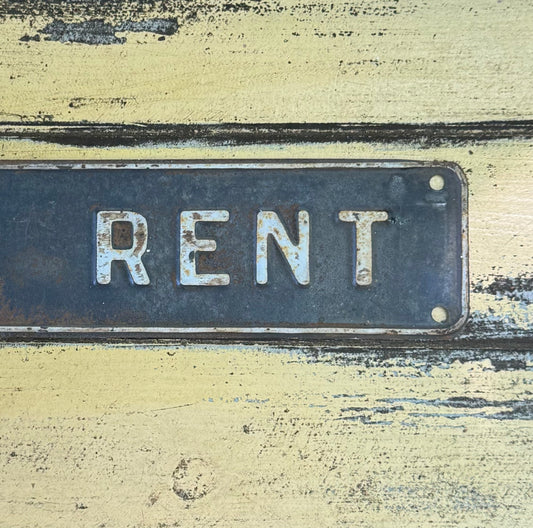 A rectangular vintage metal sign with the word 'RENT' in raised white letters, mounted on a distressed yellow wall with peeling paint.