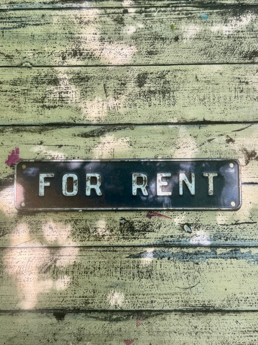 A rectangular black metal 'FOR RENT' sign with raised silver letters, mounted on a weathered green wooden plank surface.