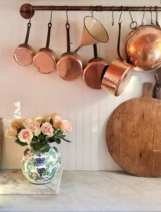 Copper pots hanging on a rack above a vase with flowers and a wooden cutting board.