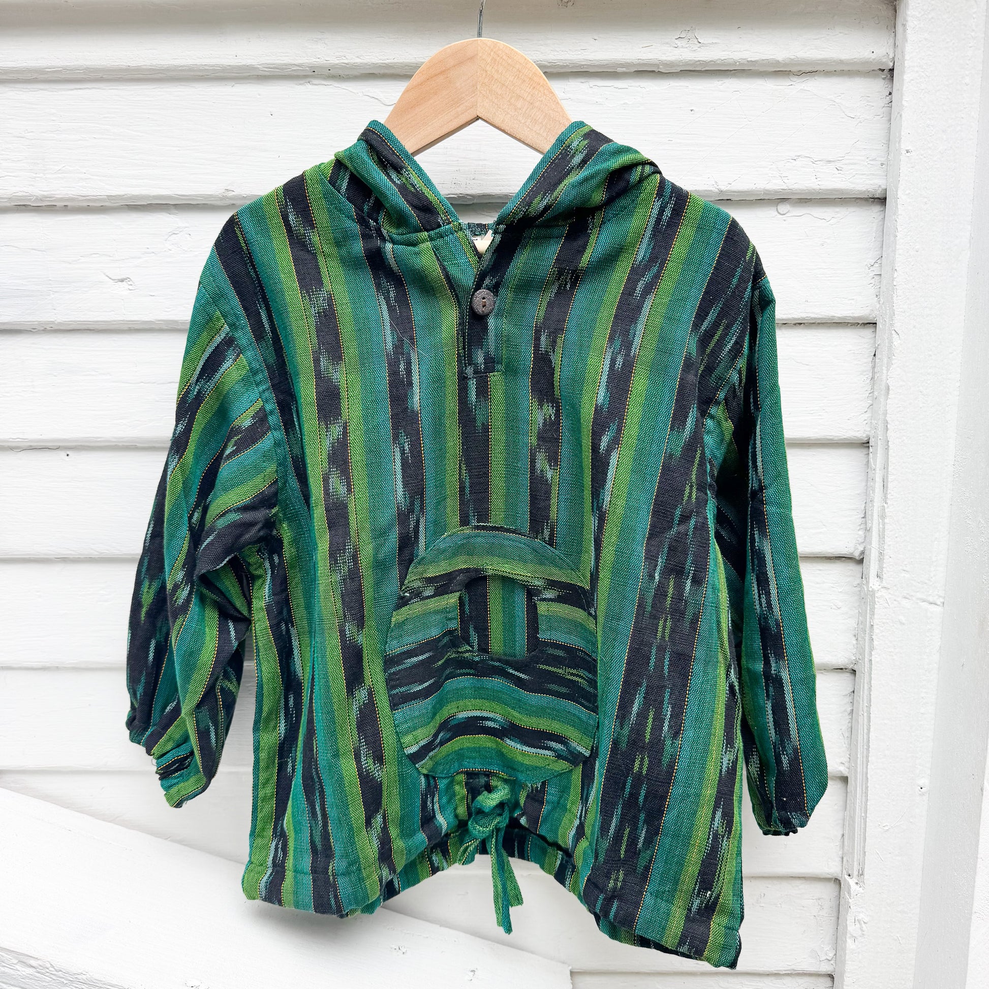 Green and black patterned poncho on a wooden hanger against a white wooden background