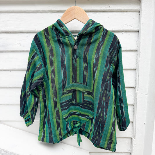 Green and black patterned poncho on a wooden hanger against a white wooden background