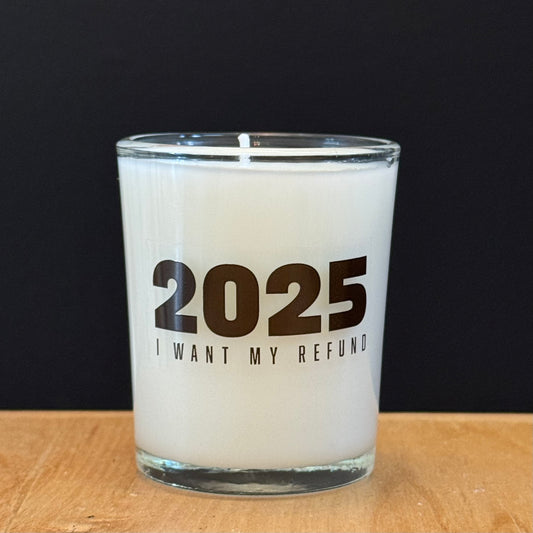 Candle with text '2025 I Want My Refund' on a wooden surface.