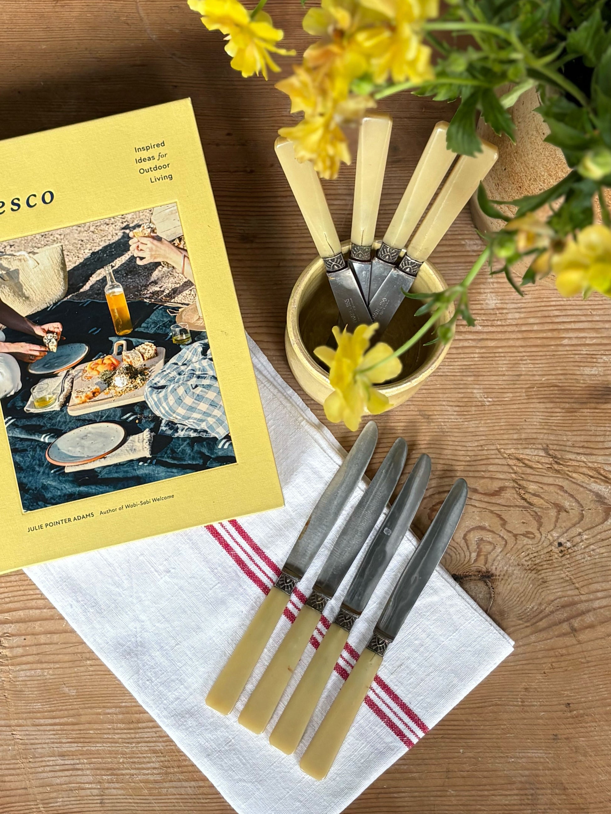 Yellow cookbook titled 'al fresco' on a wooden table with cutlery and flowers.