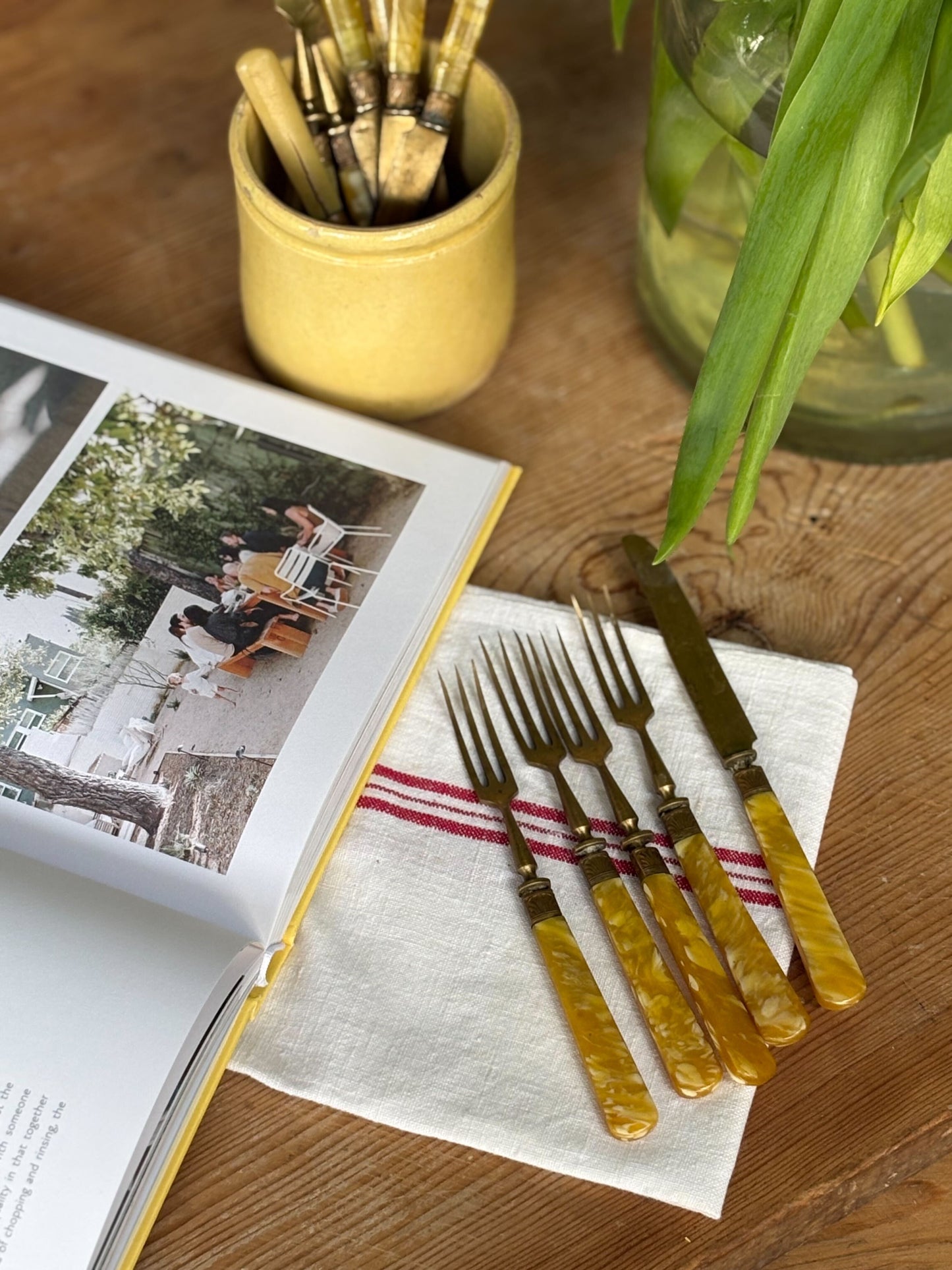 Set of yellow-handled forks and knives on a wooden table with a magazine and plant in the background.