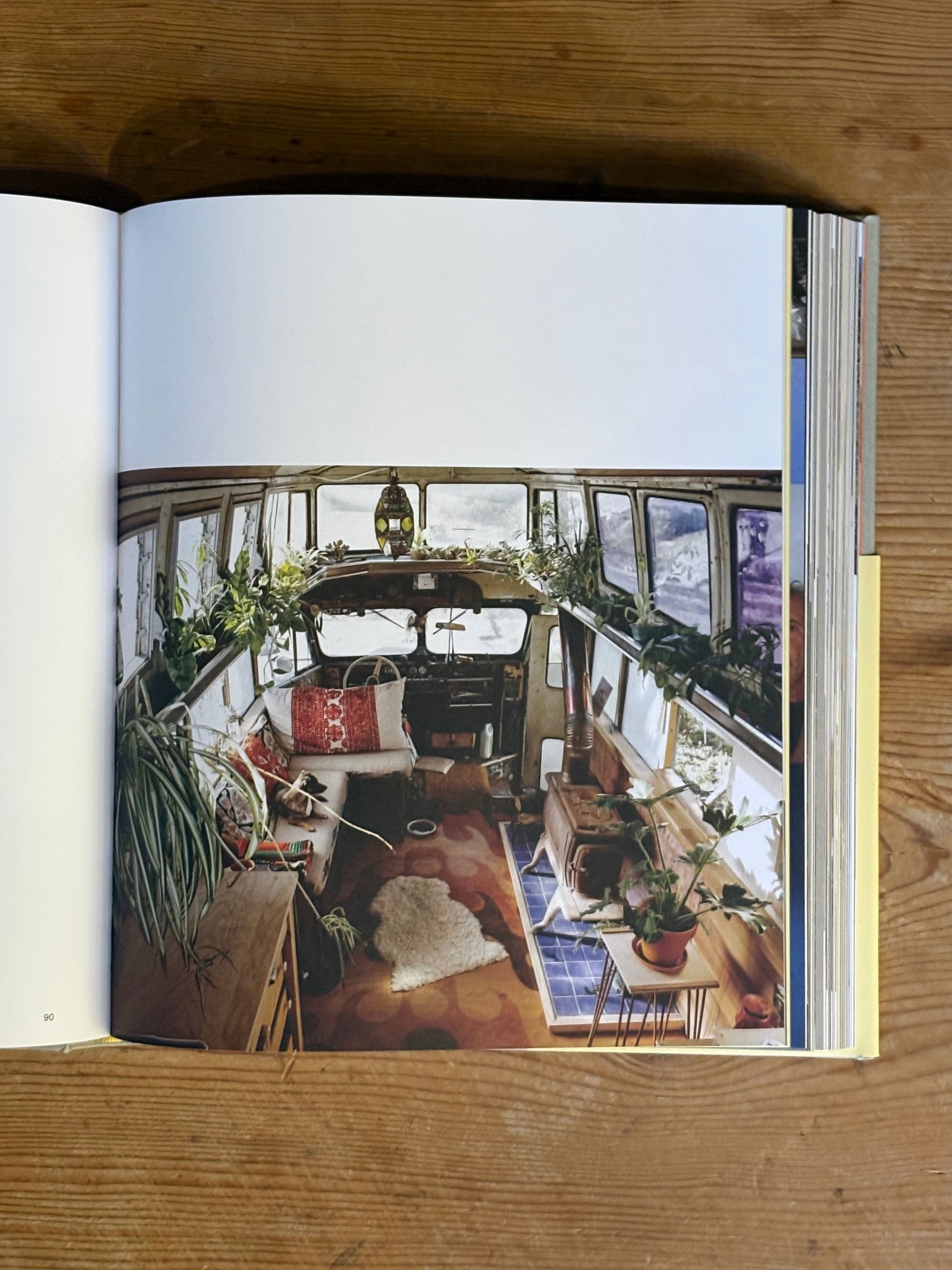 Open book showing an interior design of a converted bus with plants and furniture on a wooden surface.
