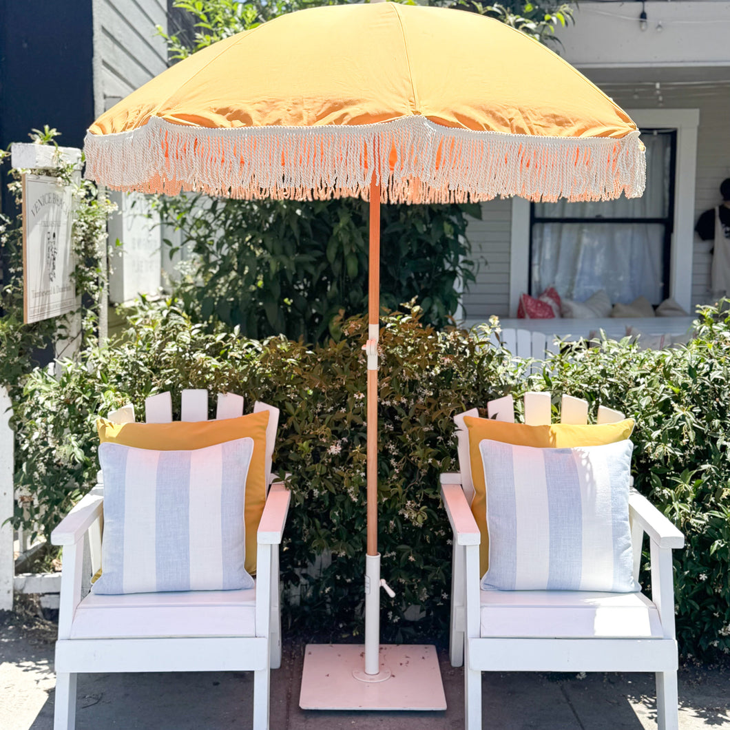 Yellow umbrella with fringe and white chairs with striped cushions on a patio.