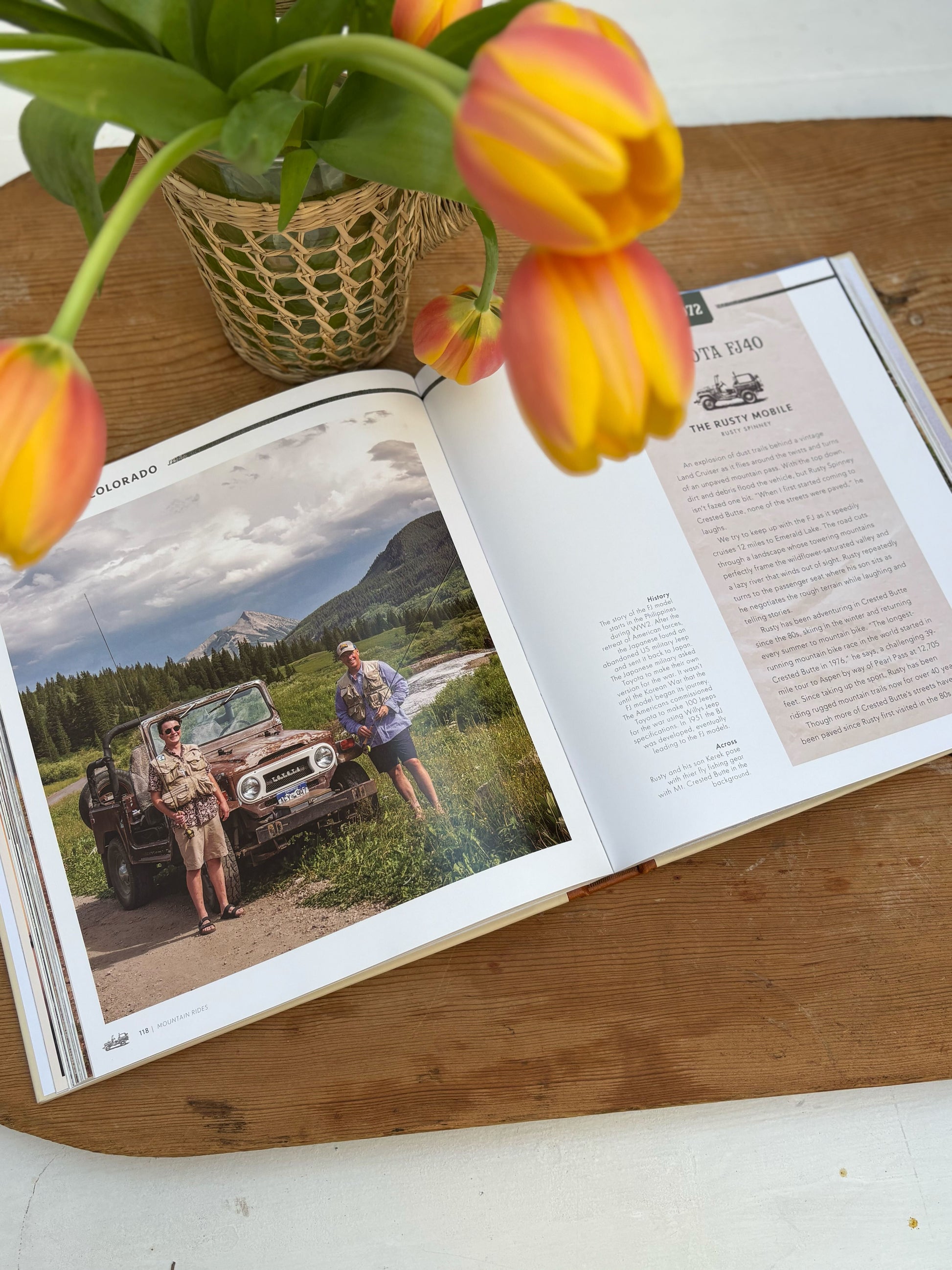 Open book on a wooden surface with a car and nature scene, surrounded by yellow and red tulips.