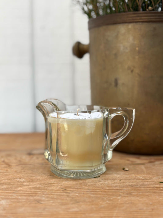Clear glass container with a candle on a wooden surface, with a blurred plant pot in the background.