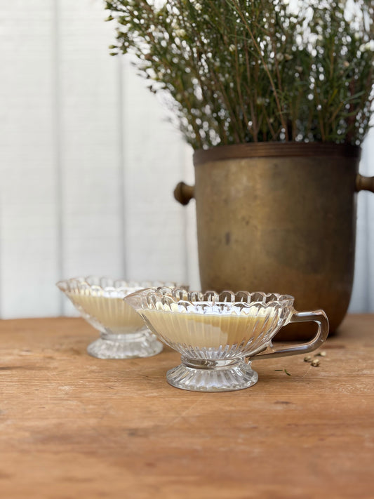 Two glass cups with handles on a wooden surface, with a potted plant in the background.
