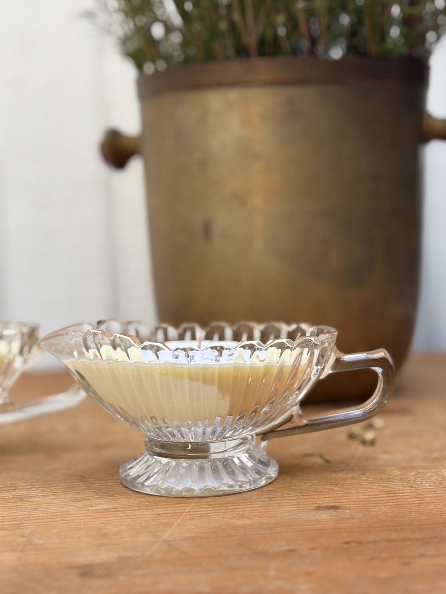 Clear glass bowl with handle on a wooden surface, with a blurred plant in the background.