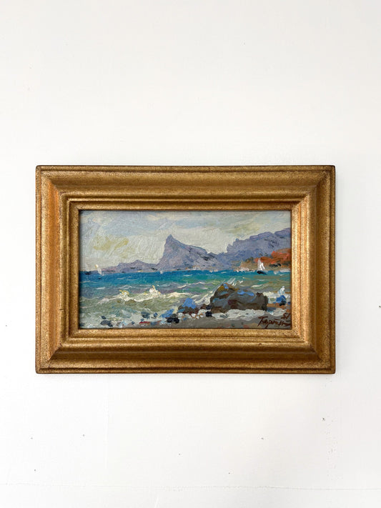 Framed painting of a coastal scene with mountains on a white wall
