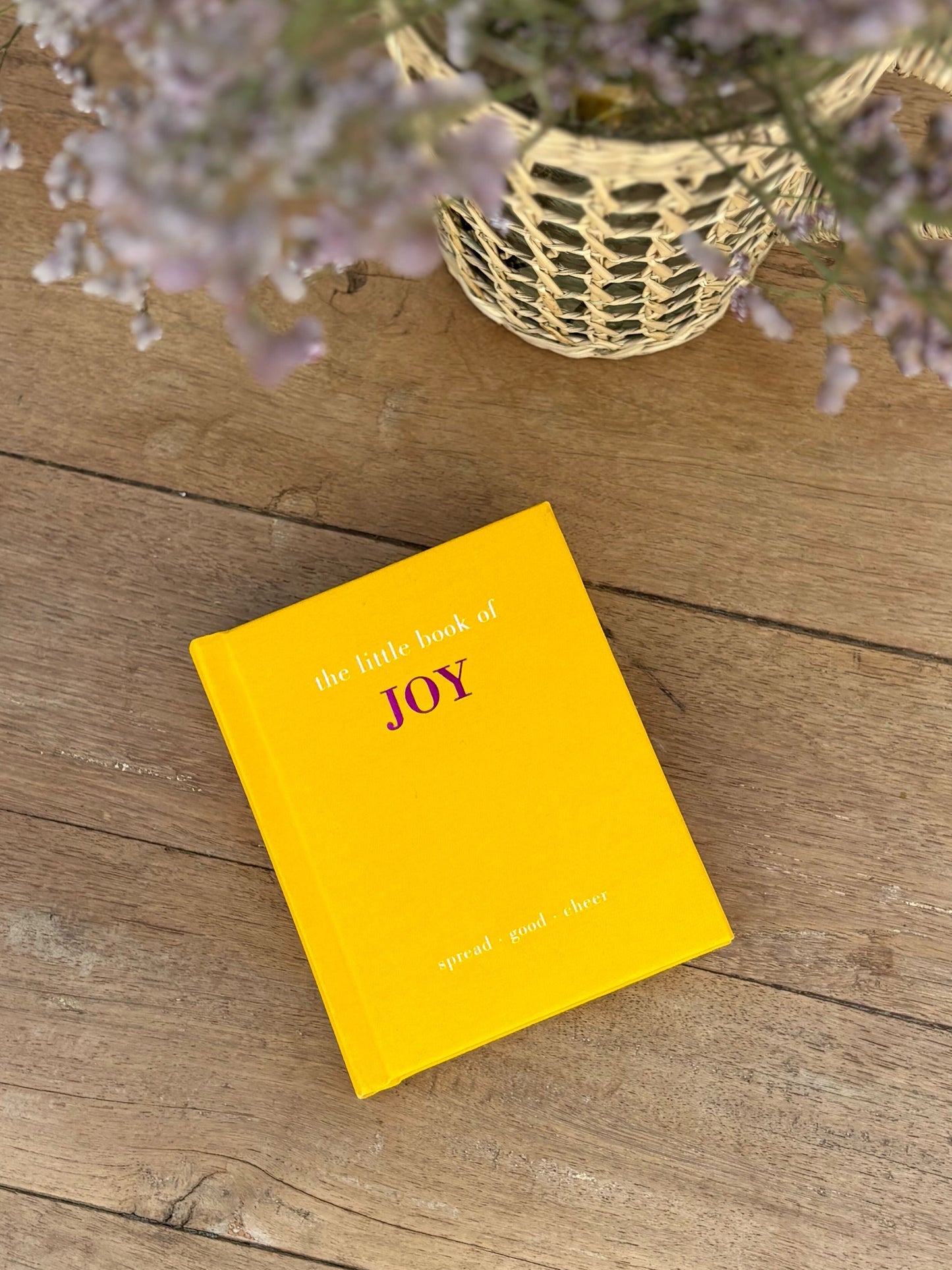 Yellow book titled 'The Little Book of Joy' on a wooden surface with a basket of flowers.