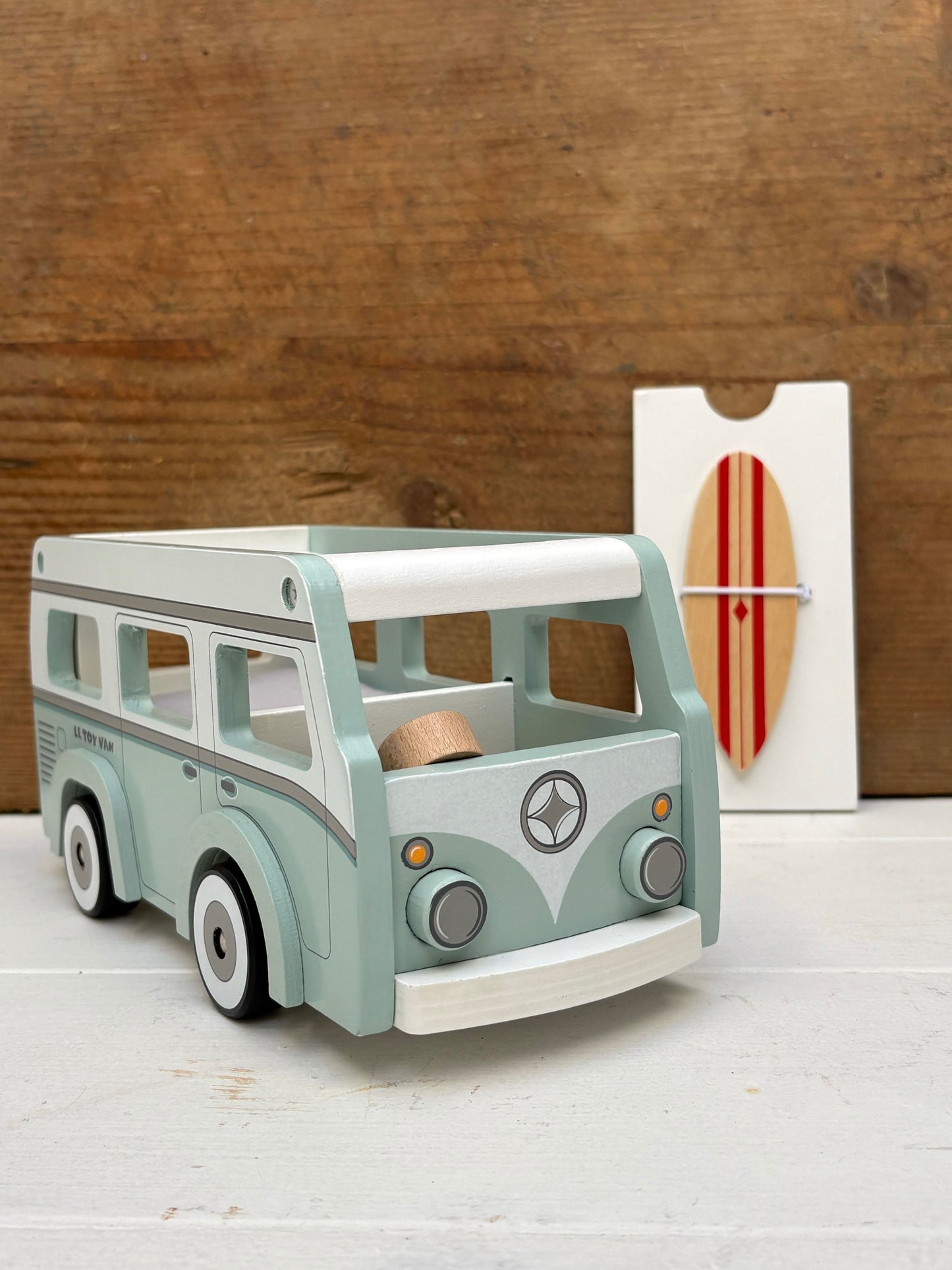 Wooden toy van on a white surface with a wooden wall and surfboard in the background