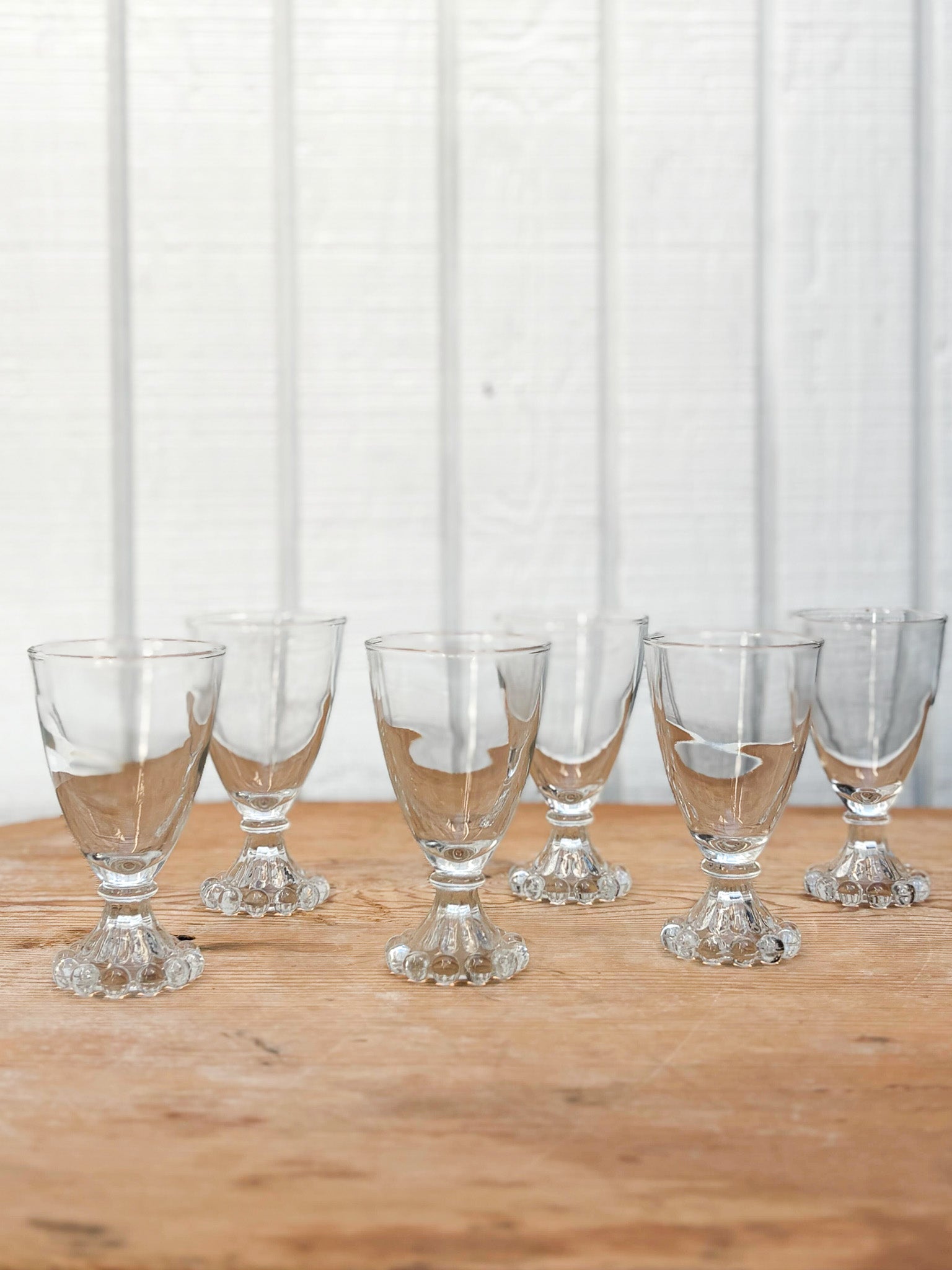 Set of six clear glass goblets on a wooden surface with a white curtain background