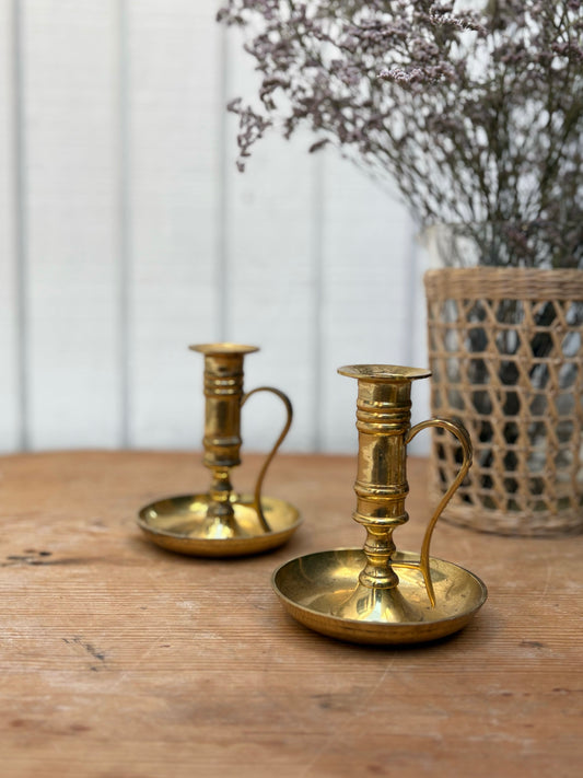 Two brass candle holders on a wooden surface with a white wall and basket in the background.