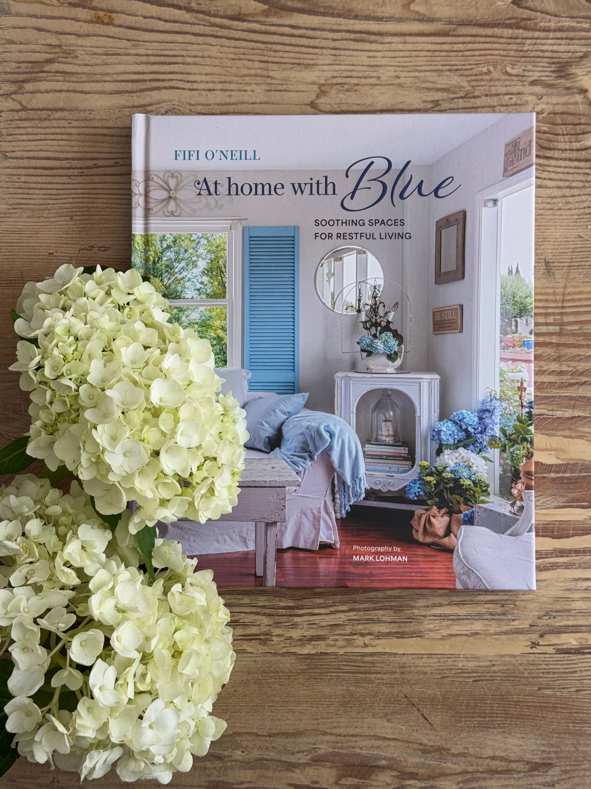 Book titled 'At Home with Blue' by Fifi O'Neill on a wooden surface with hydrangeas.