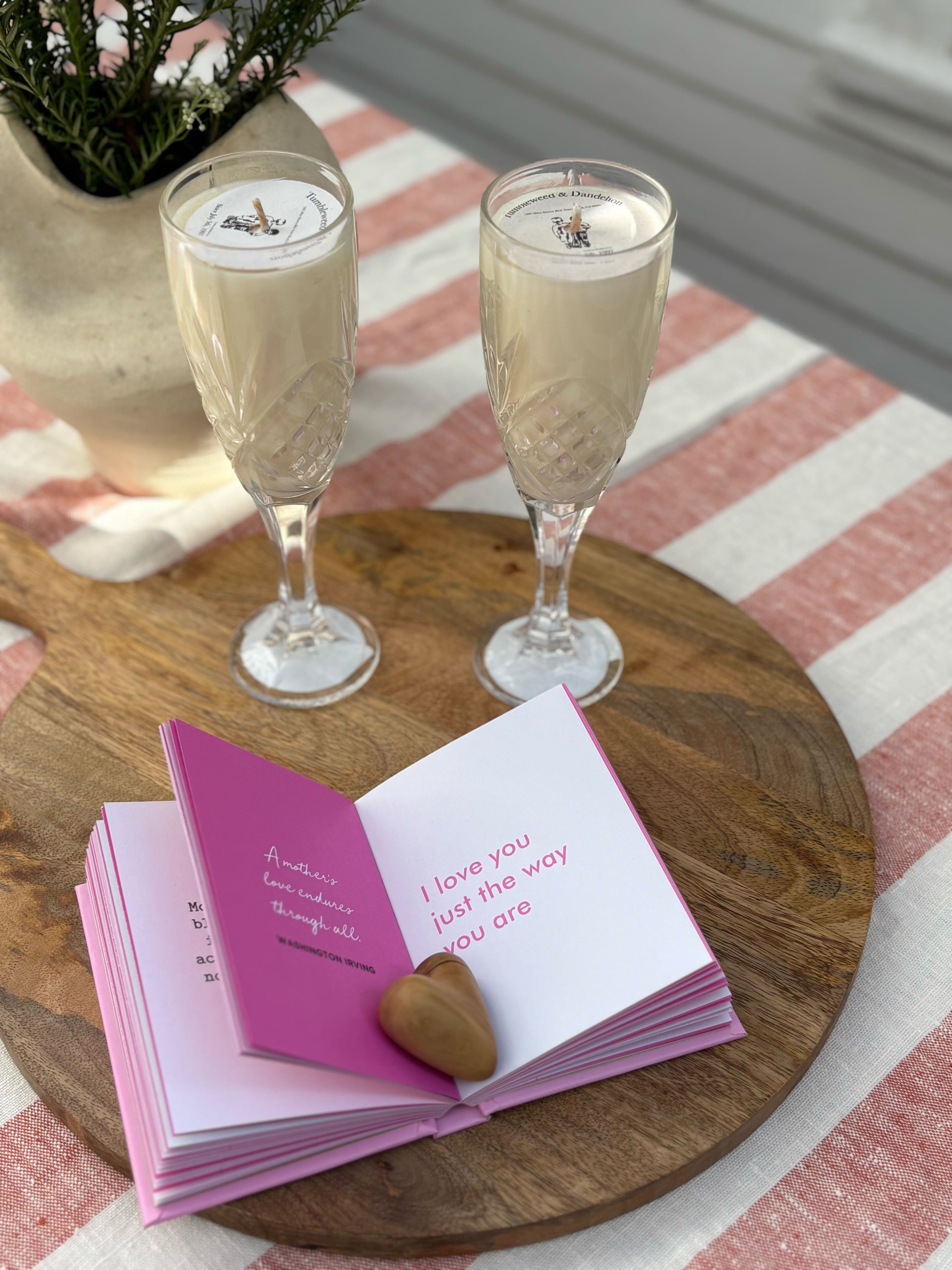 Two champagne glasses with candles in them with a small book and heart-shaped object on a wooden tray.