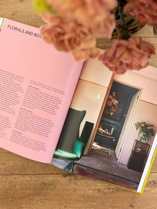 Open book with a pink page showing text and a photo of green boots, with a vase of flowers on top.