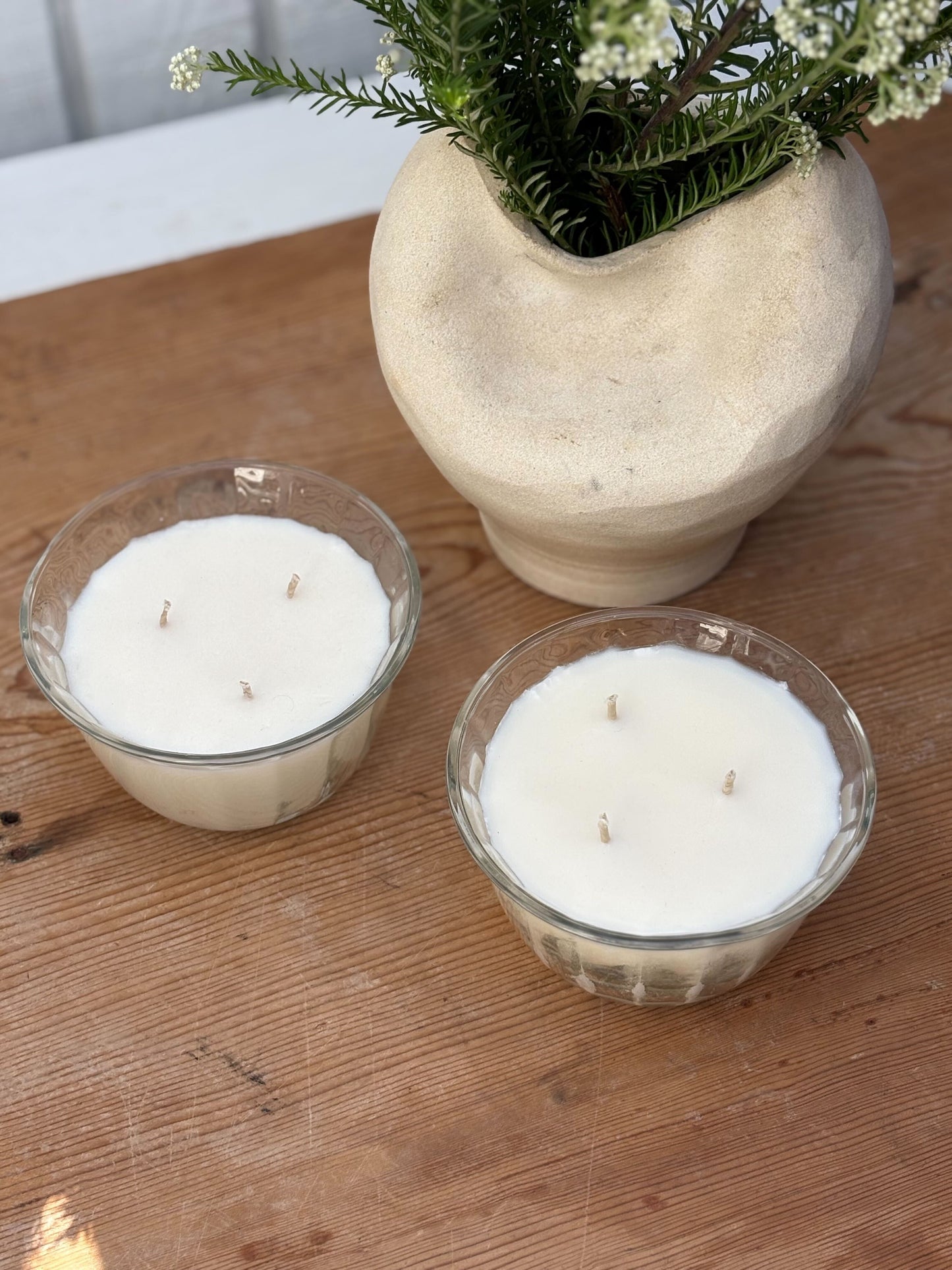 Two glass candles with a white candle inside on a wooden surface, accompanied by a decorative vase with greenery.