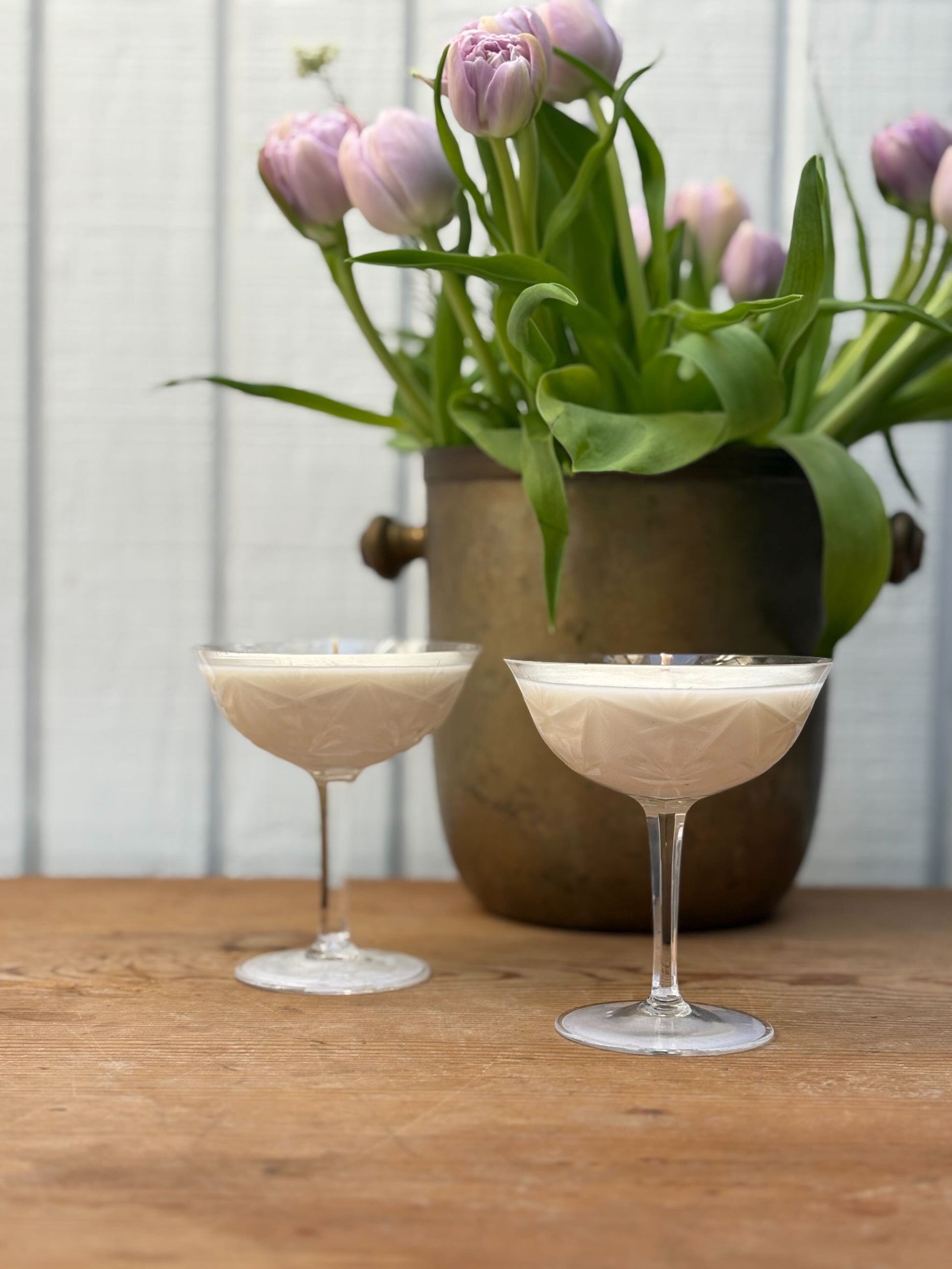 Two cocktail glasses with a creamy beverage on a wooden table, with a vase of tulips in the background.