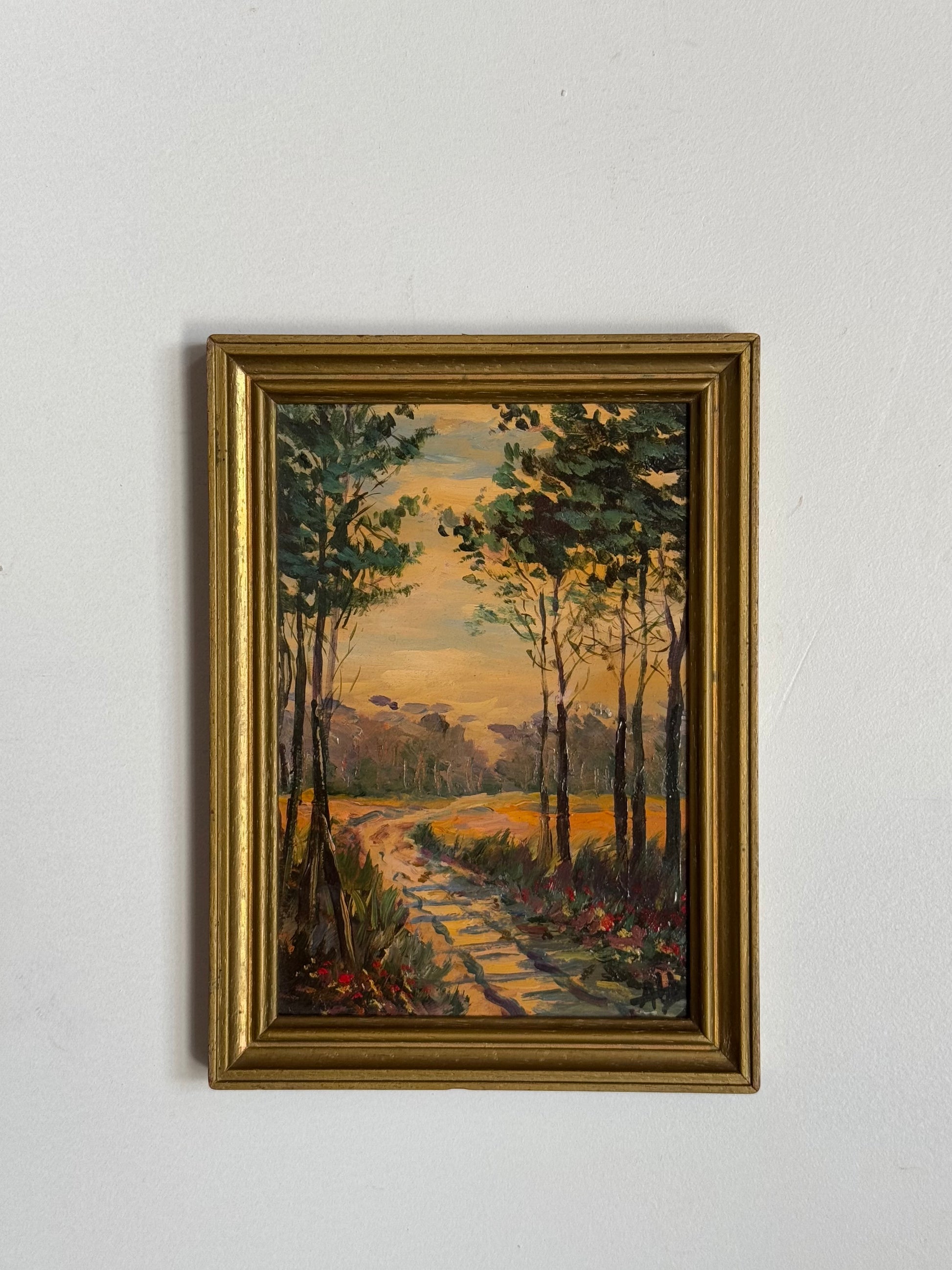 Framed painting of a forest path with a sunset sky on a white wall.