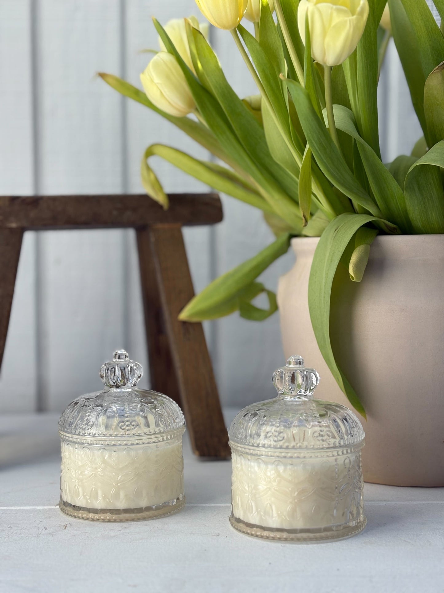 Two glass jars with decorative lids on a white surface with a vase of yellow tulips in the background.