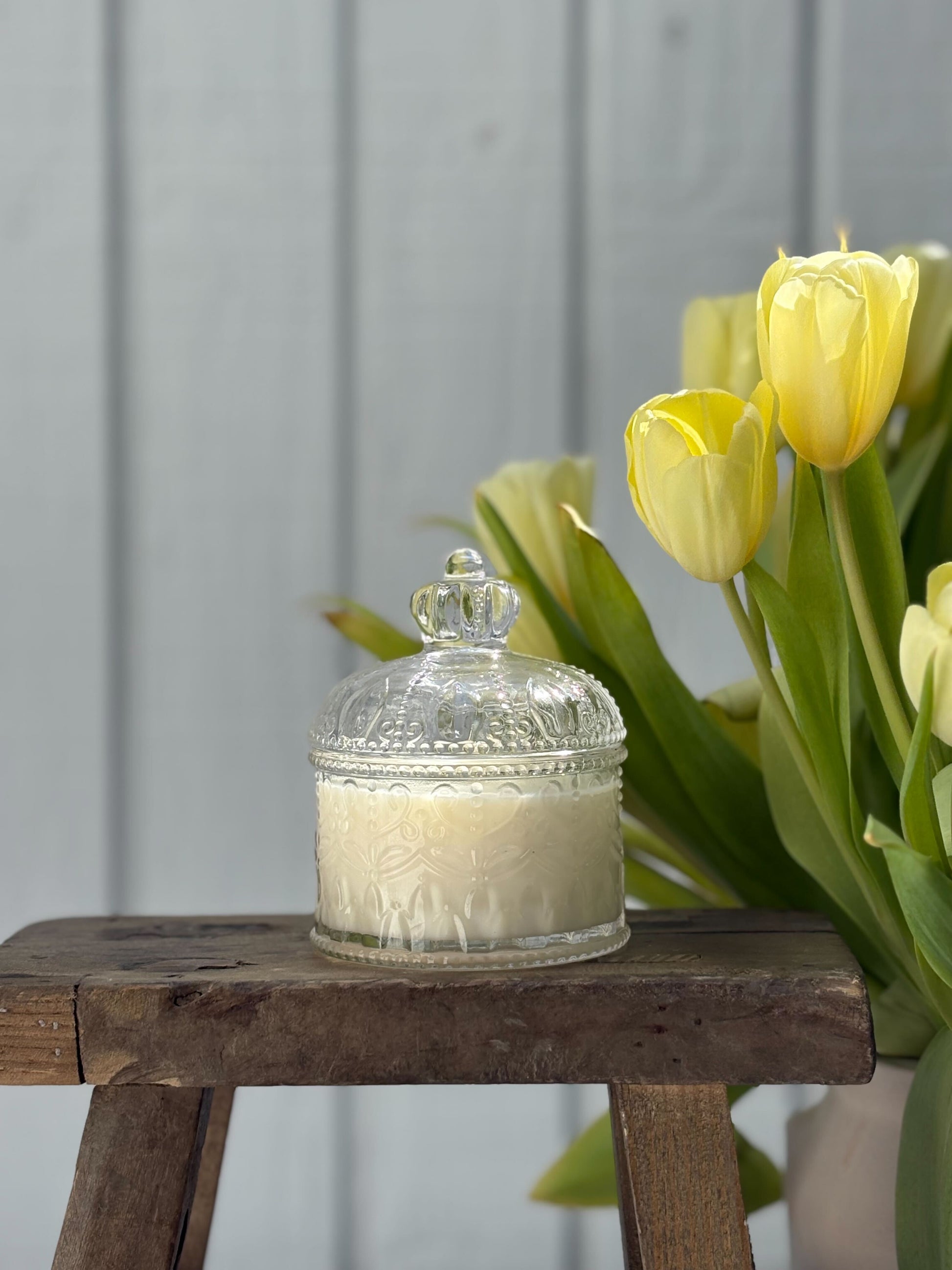 Decorative candle in a glass holder on a wooden stool with yellow tulips in the background.