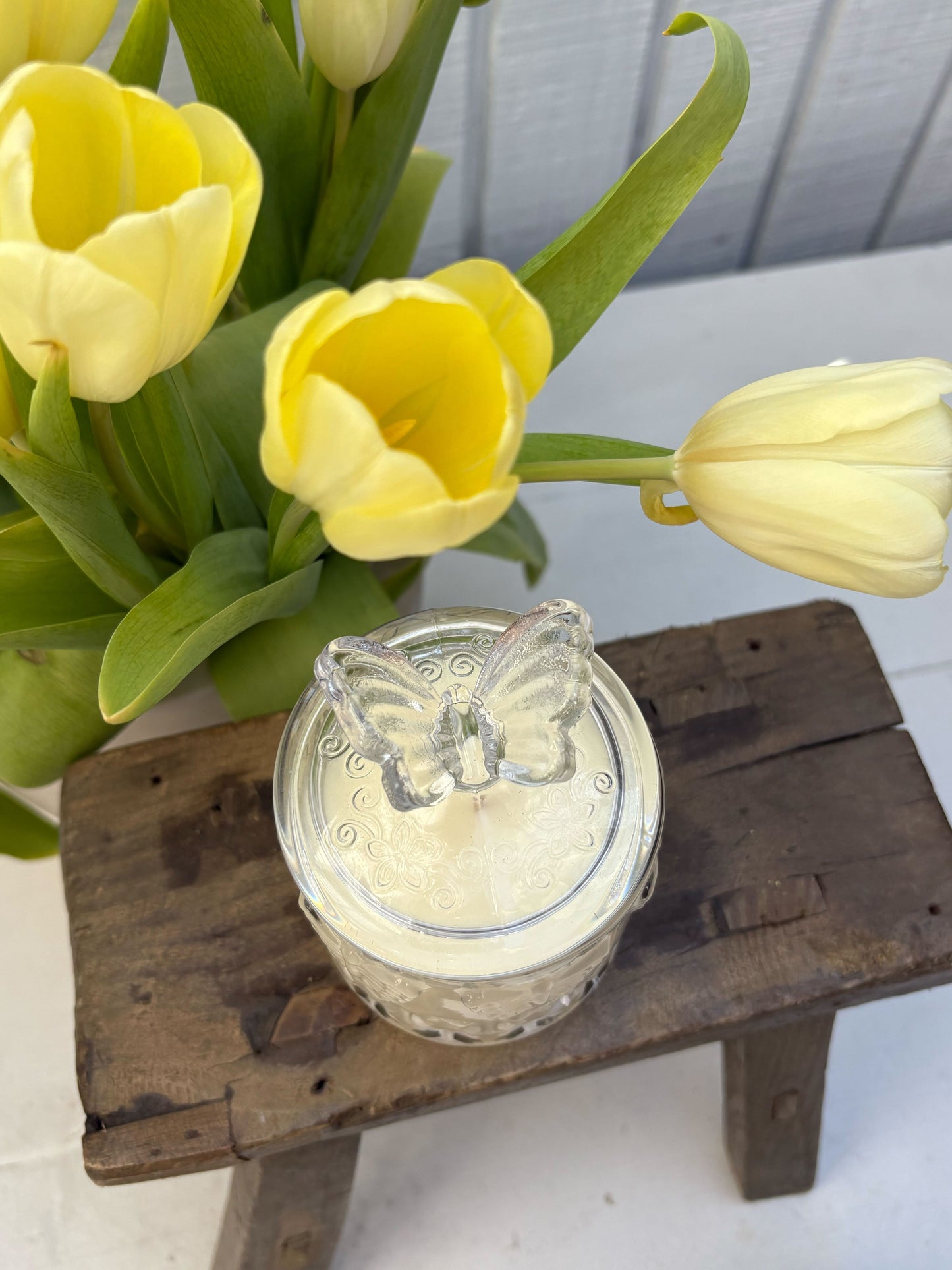 Clear glass jar with butterfly design lid on a wooden stand, surrounded by yellow tulips.