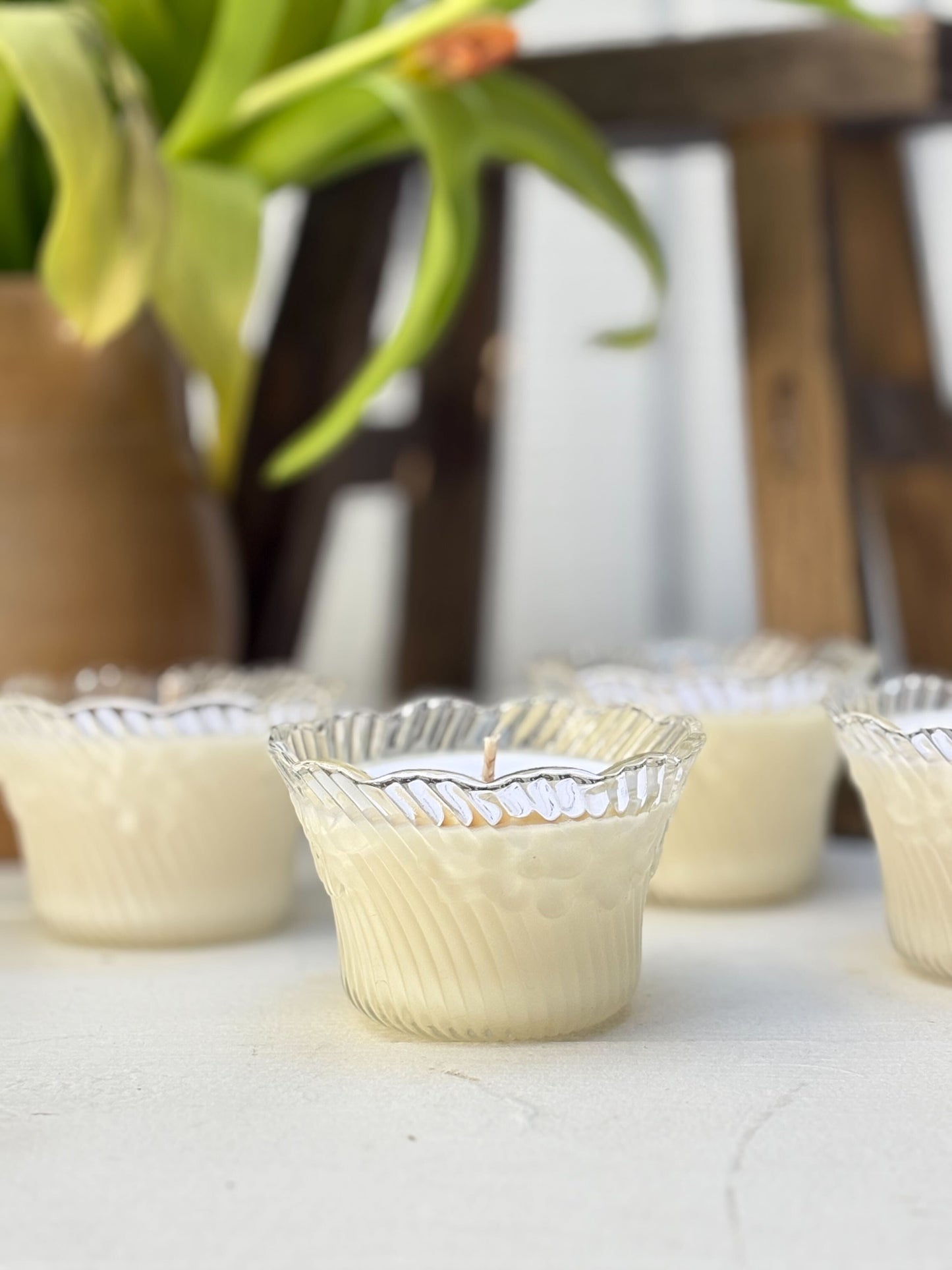 Row of small candles in decorative holders on a light surface with a blurred plant and wooden stool in the background.