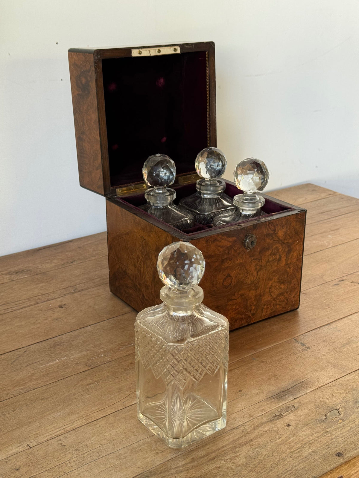 A clear cut-glass decanter bottle with a faceted stopper sits in front of an open dark wood presentation box containing three similar glass decanters with round crystal-style stoppers, placed on a wooden tabletop against a plain light wall.