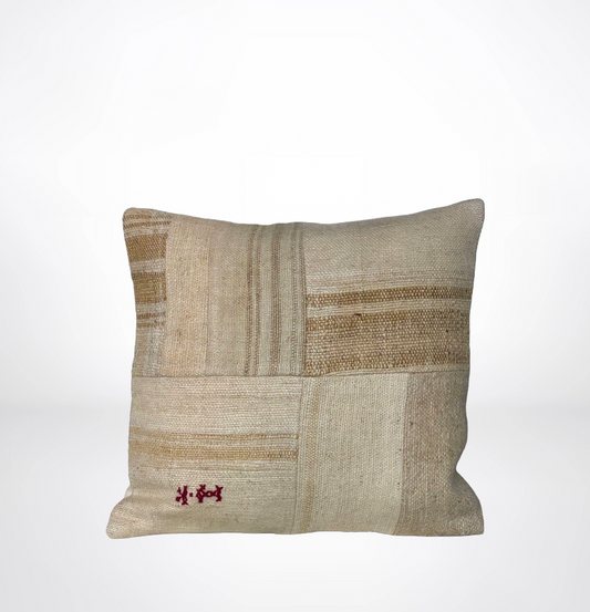 A square decorative throw pillow with a patchwork design of natural linen and woven beige panels, featuring a small red embroidered motif in the lower left corner, set against a plain white background.