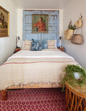Load image into Gallery viewer, small cottage bedroom with bed, vintage doors as headboard painted in sky blue and distressed, vintage paintings and baskets as decor, red rug with small diamond pattern