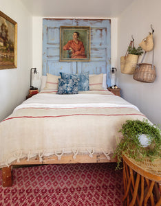 small cottage bedroom with bed, vintage doors as headboard painted in sky blue and distressed, vintage paintings and baskets as decor, red rug with small diamond pattern