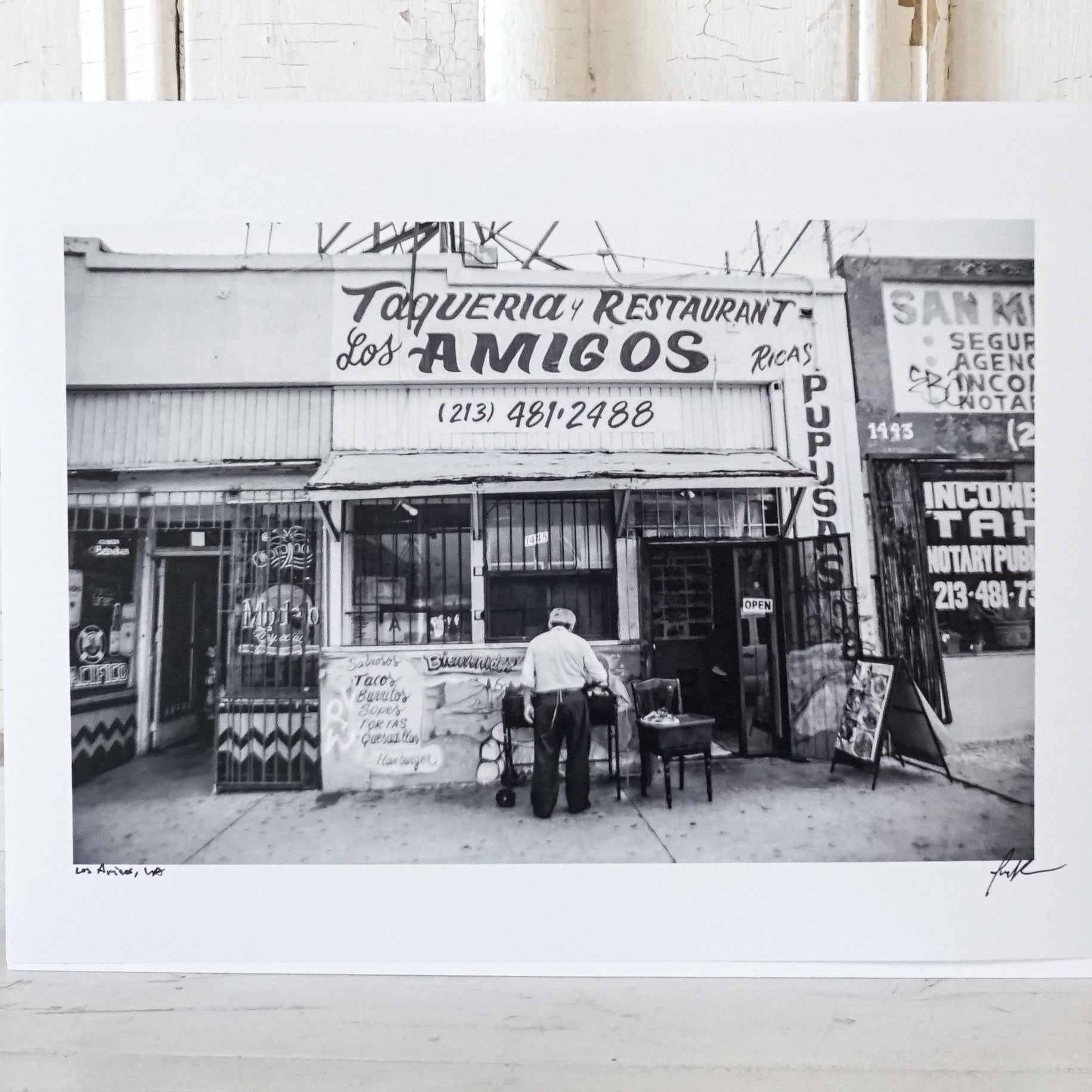 black and white photo of front of an old taqueria with man standing in front  Edit alt text