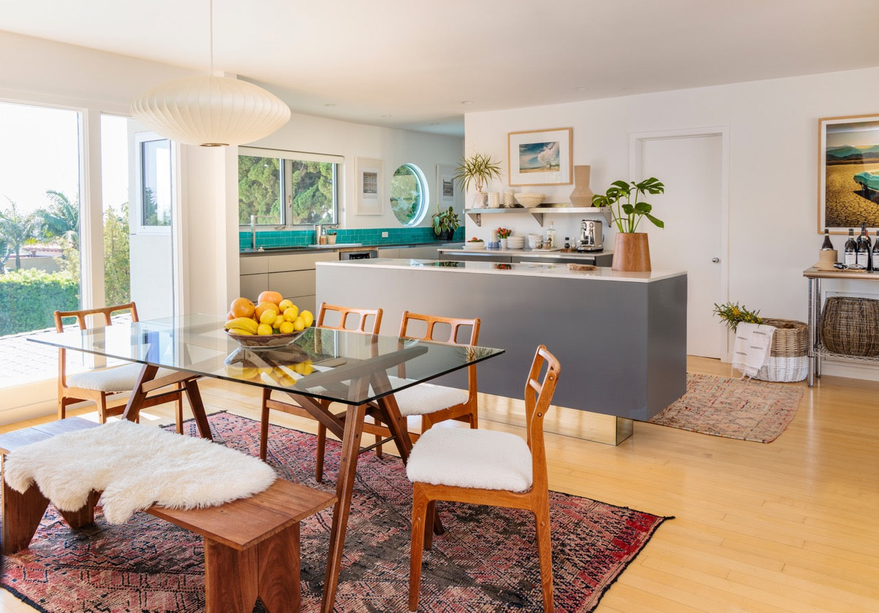 dining are of a home with wood and glass dining table, wood chairs with upholstered seats, wood bench with sheepskin rug on seat, gray colored bar area  with shelves behind it and artwork