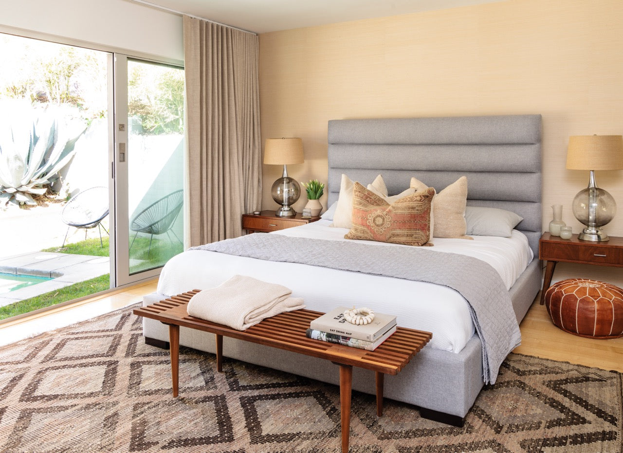 Gray upholstered bed with diamond patterned tan and brown rug, brown end table at foot of bed, leather pouf, brown wood nightstands with glass lamps
