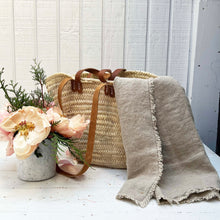 Load image into Gallery viewer, natural colored linen throw blanket with rustic frayed edges, hanging out of a wicker market basket with leather handles