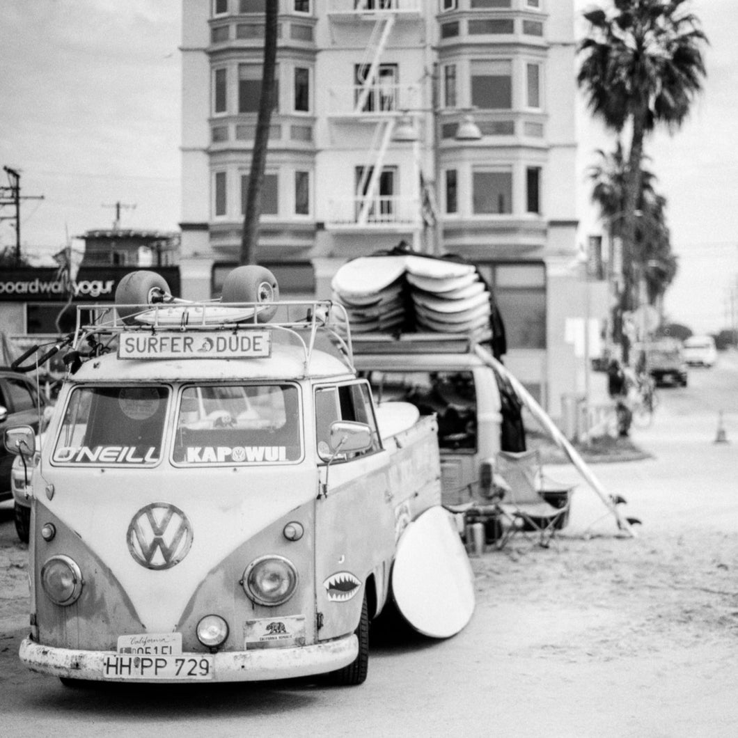 black and white photograph of a vintage VW van, stacks of surfboards and building with palm trees