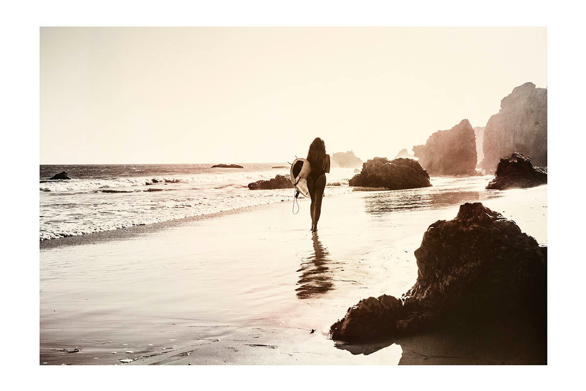 sepia toned photograph of woman with surfboard on beach with rocks  Edit alt text
