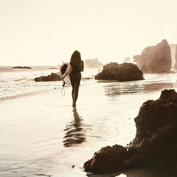 sepia toned photograph of woman with surfboard on beach with rocks