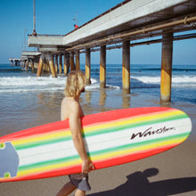 Load image into Gallery viewer, photo of blonde hair man holding surfboard walking on beach next to a pier