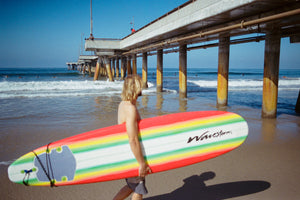 photo of blonde hair man holding surfboard walking on beach next to a pier